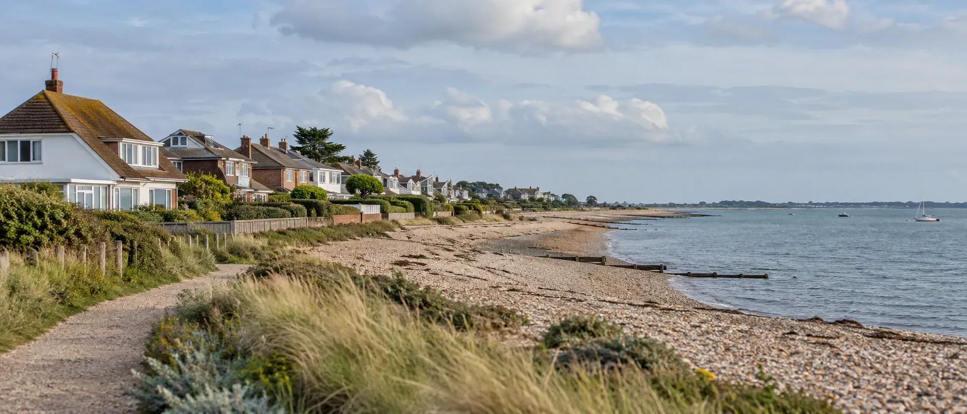 Illustrative view of coastal homes beside a shingle beach and footpath under a cloudy sky.