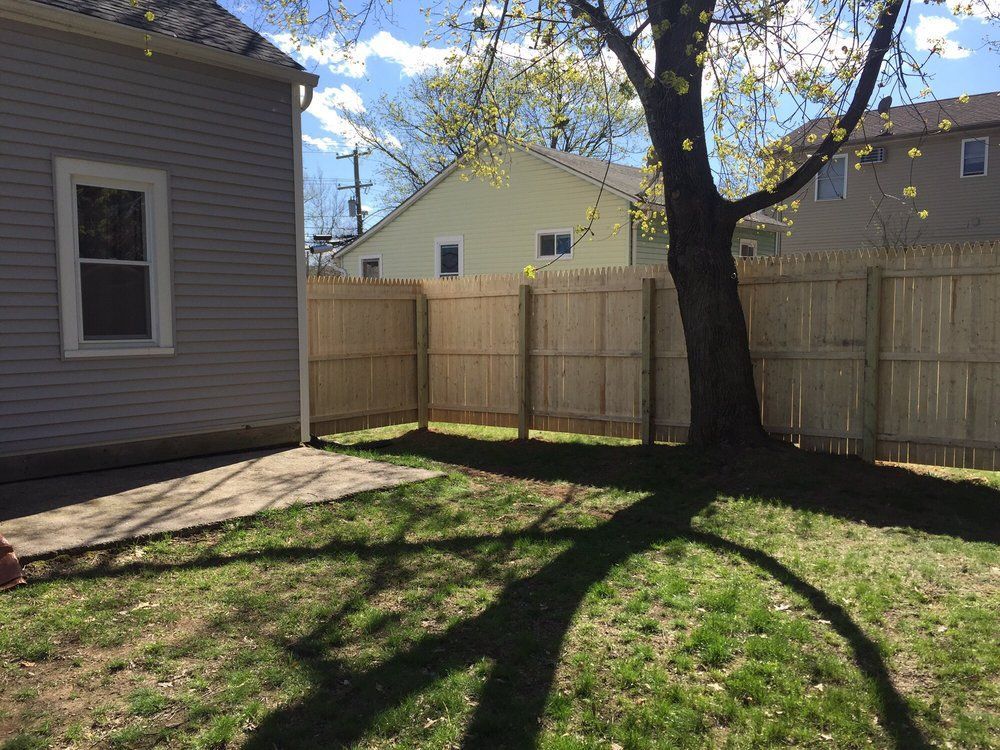 A backyard with a wooden fence and tree, cast shadow on grass, adjacent to a house with a window.