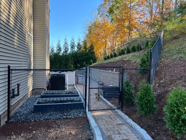 Black chain-link fence with gate, garden beds, and stone path next to a house and hillside.
