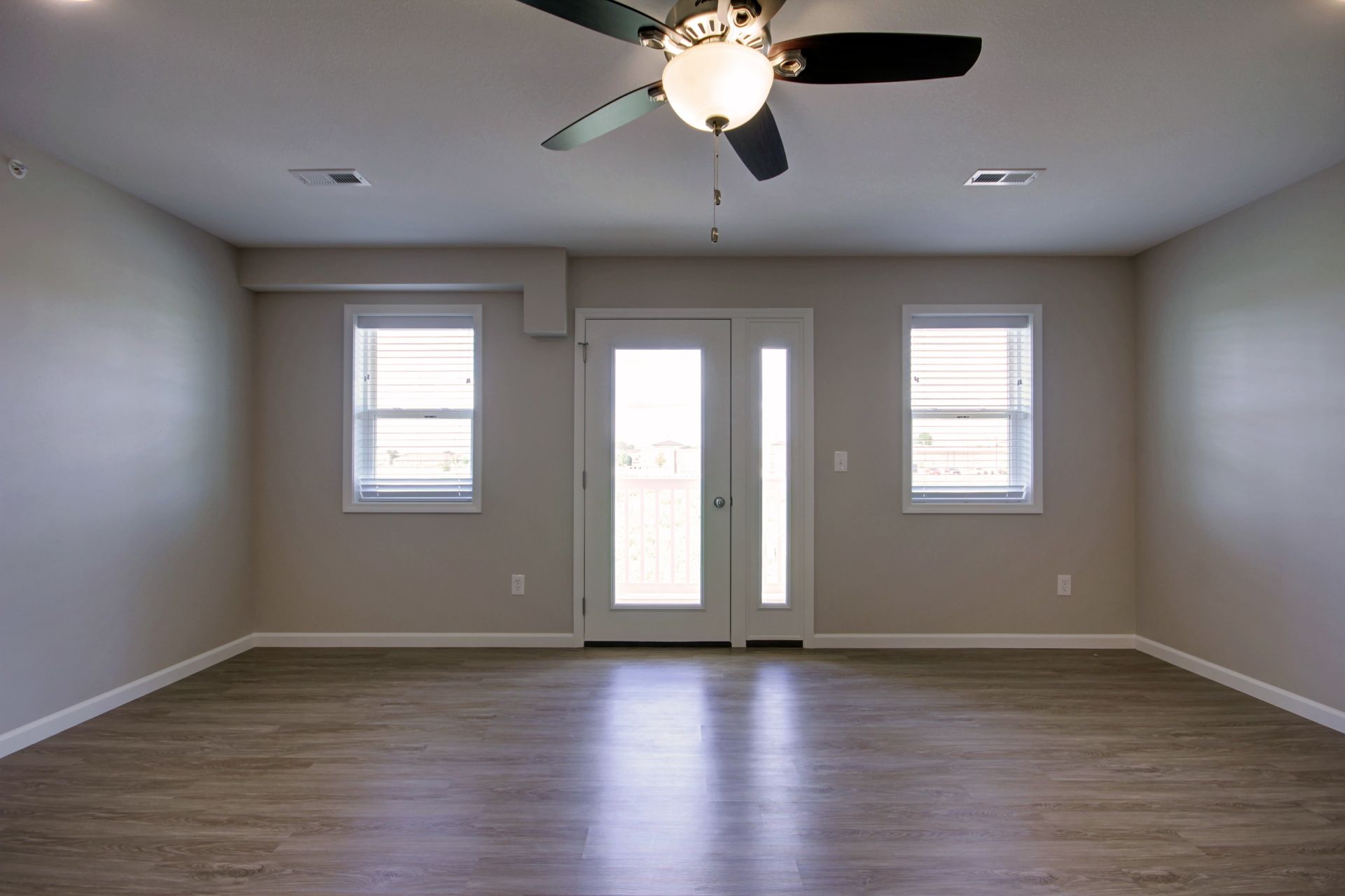Empty living room with gray walls, wood floor, ceiling fan, and double glass doors flanked by two windows
