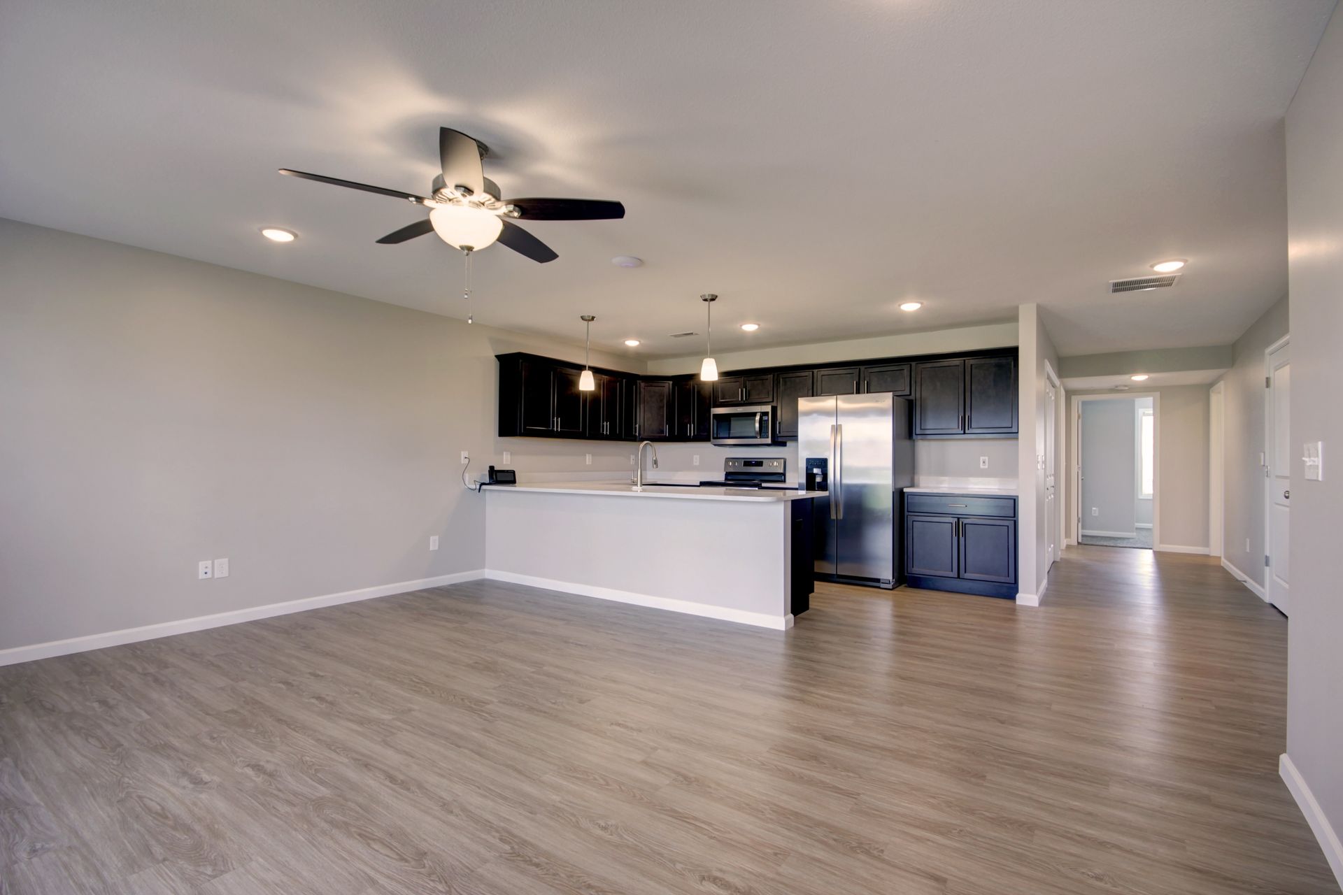 Empty open-plan living room with wood floors, ceiling fan, and dark kitchen cabinets by a hallway