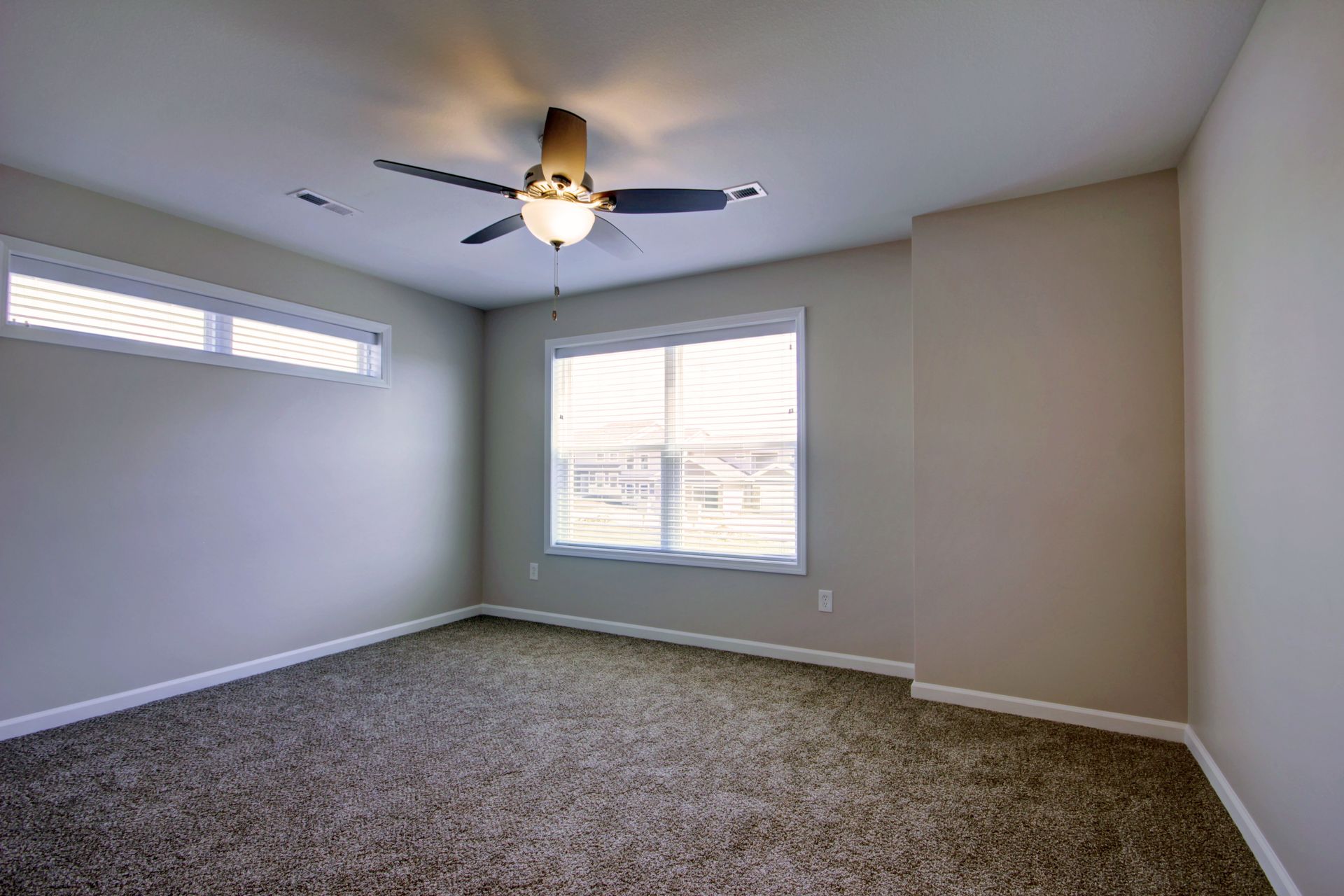 Empty gray bedroom with carpet, ceiling fan, and two windows letting in daylight