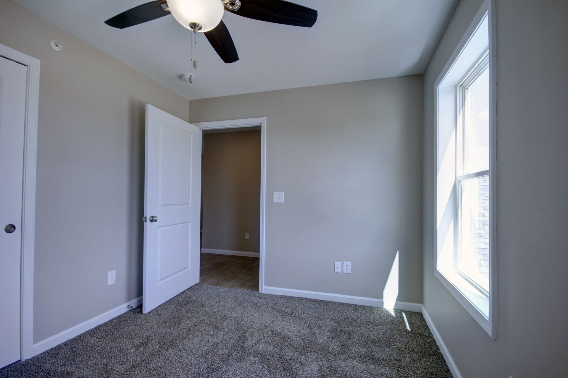 Empty gray bedroom with carpet, ceiling fan, open door, and large window letting in daylight