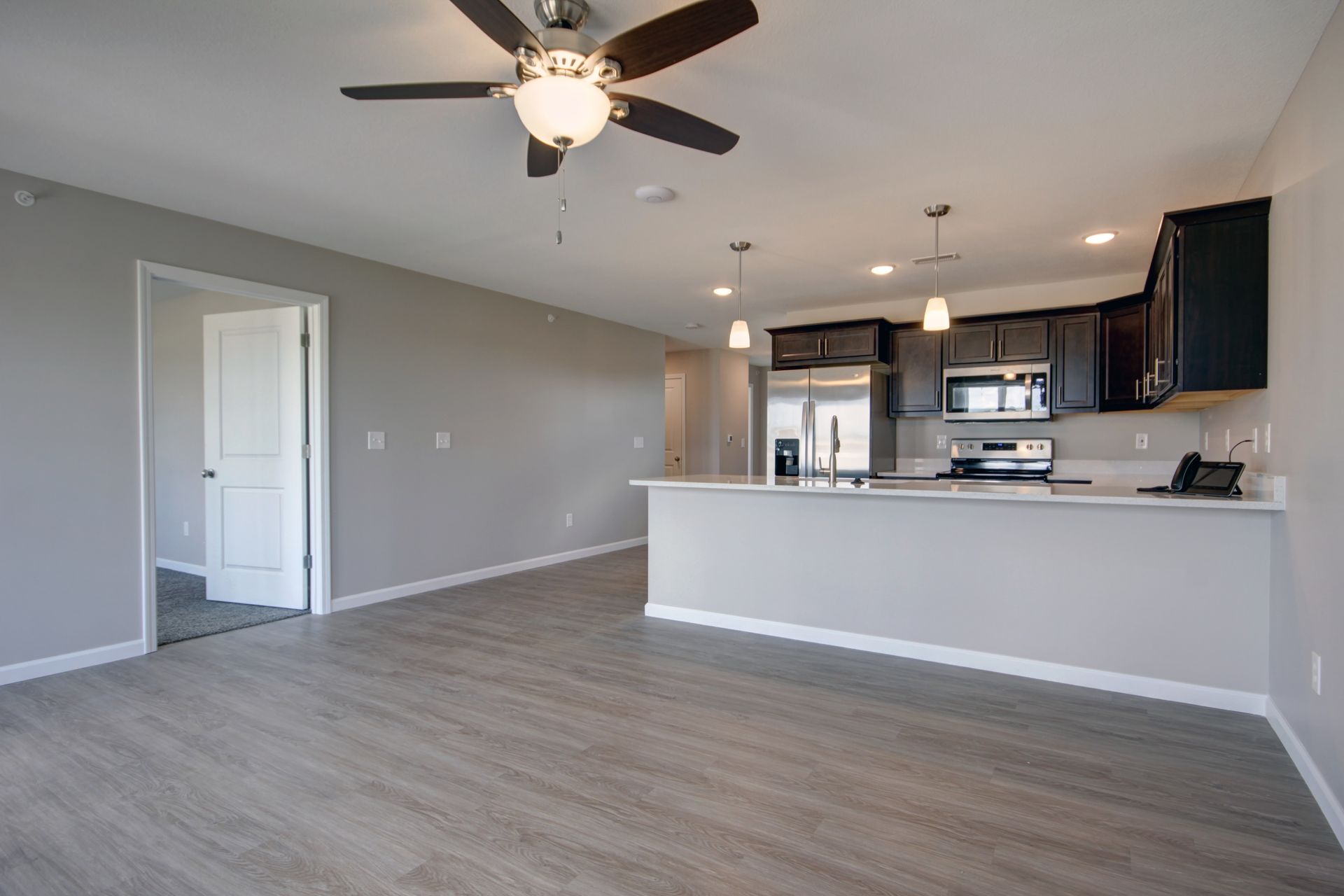 Open-plan living room with gray floors, ceiling fan, and a kitchen with dark cabinets and white island.