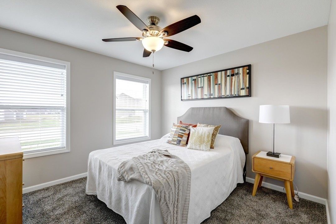 Bright bedroom with a bed, nightstand, lamp, ceiling fan, windows, and gray carpet