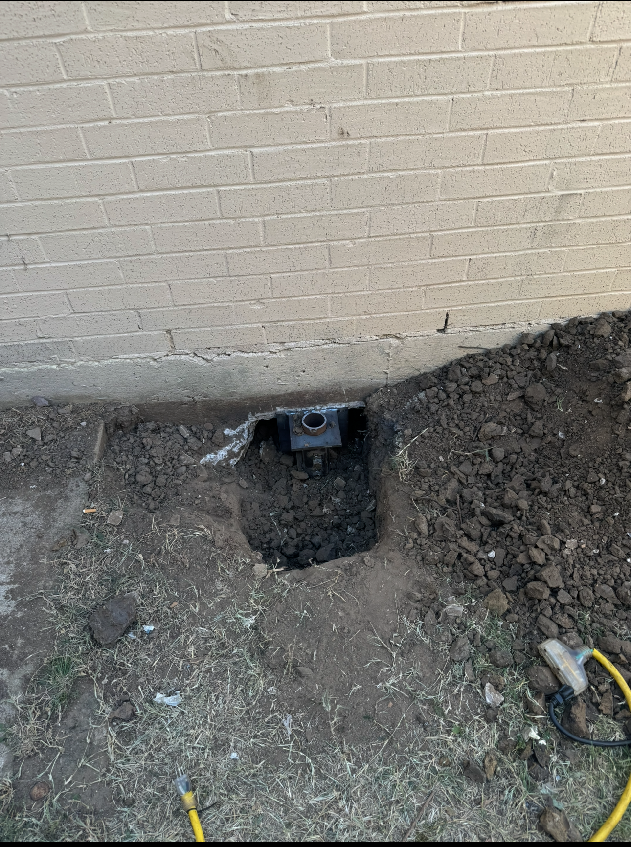 Hole dug near brick wall, revealing a utility box. Dark soil surrounds the hole, a shovel and yellow hose visible.