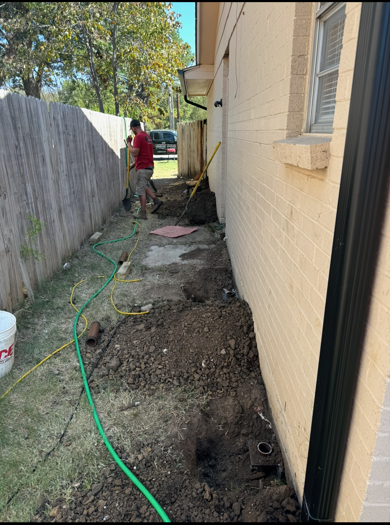 Man digging a trench along a brick building and wooden fence. Green hose and bucket present.