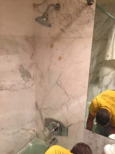 Bathroom shower with marble tiles, wall-mounted showerhead and faucet, and a person bending near a mirror.
