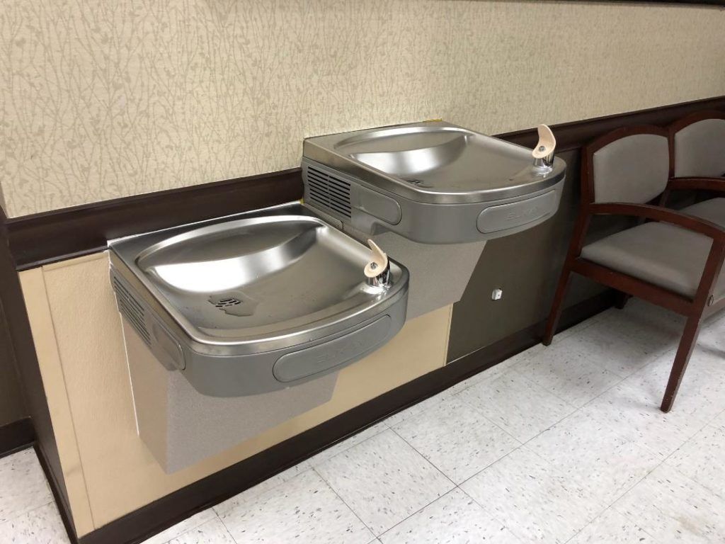 Two stainless steel drinking fountains mounted on a beige wall beside a chair