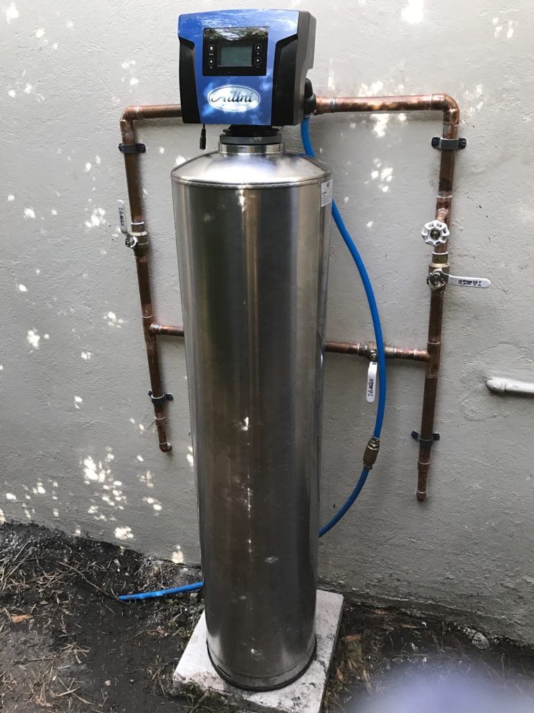 Tall stainless steel water softener tank with control head and blue hose against a basement wall