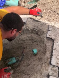Worker leaning into a dug-out pit with gray soil and broken concrete debris, wearing red gloves and glasses
