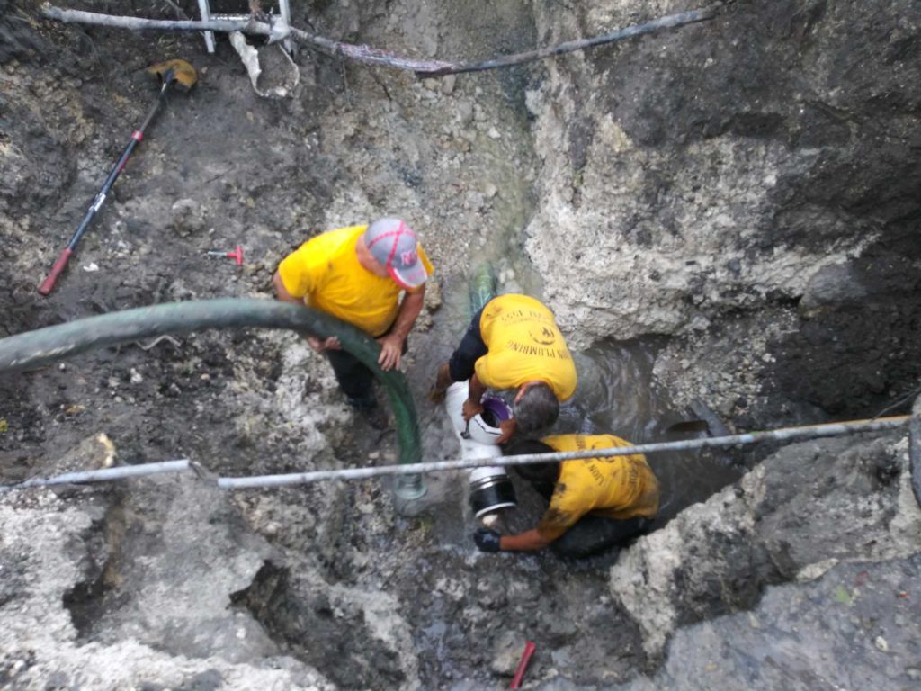 Three workers in yellow shirts digging in a rocky trench beside large hoses and pipes.