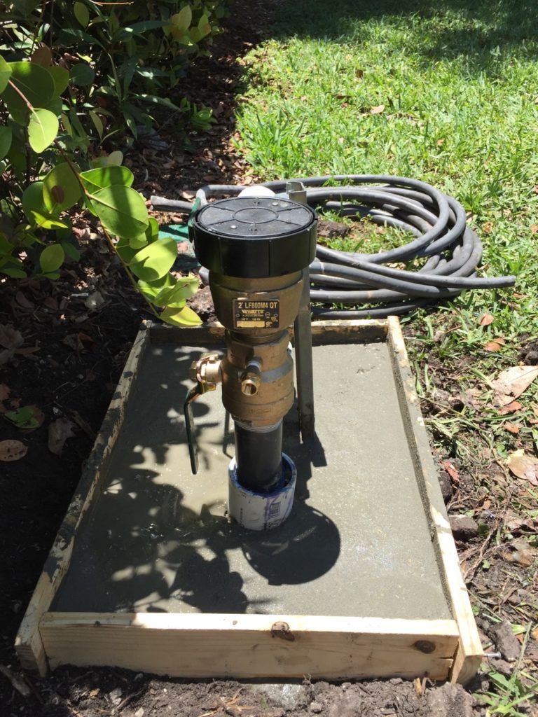 Backyard water pump in a concrete box beside coiled black hoses and green grass
