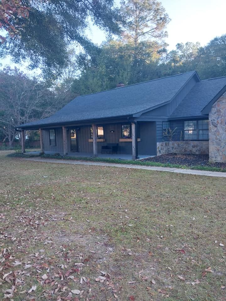 Low, dark-roofed building with porch. Gray walls, stone accent, windows, and short grass in front.