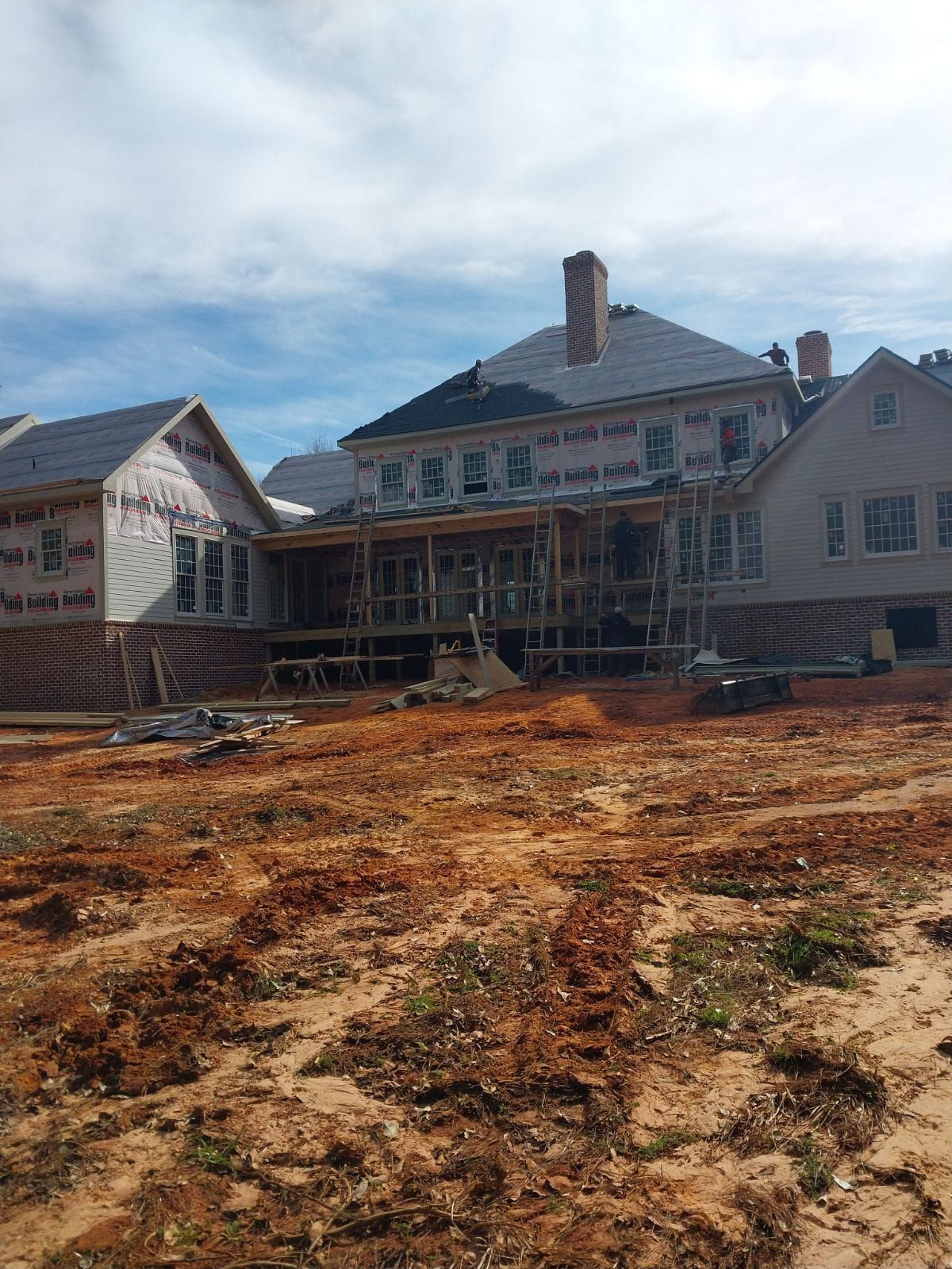 A house under construction with exposed wood framing, covered in building paper, and surrounded by dirt.