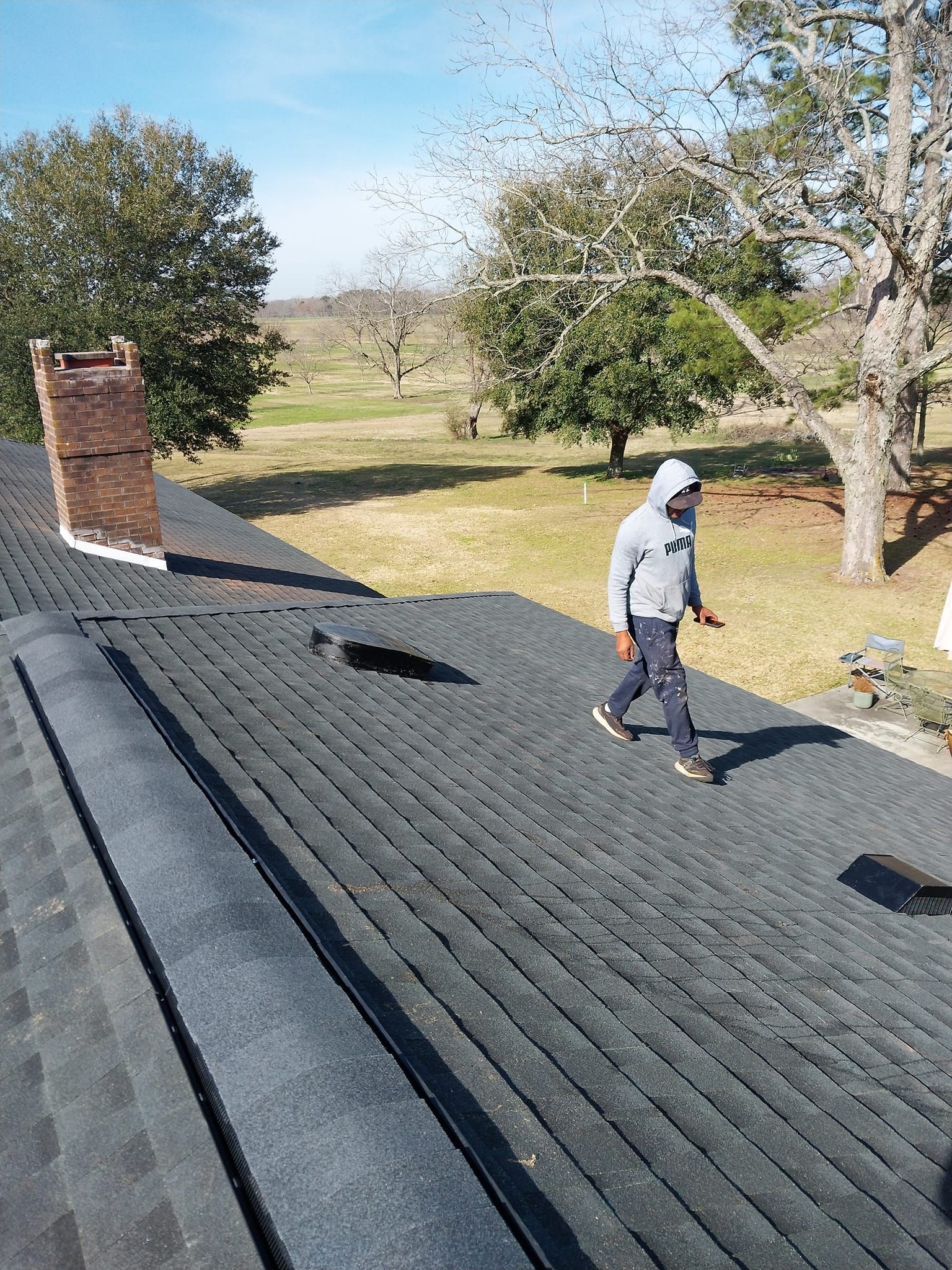 Person in hoodie walking on a dark shingled roof near a brick chimney and trees in a field.
