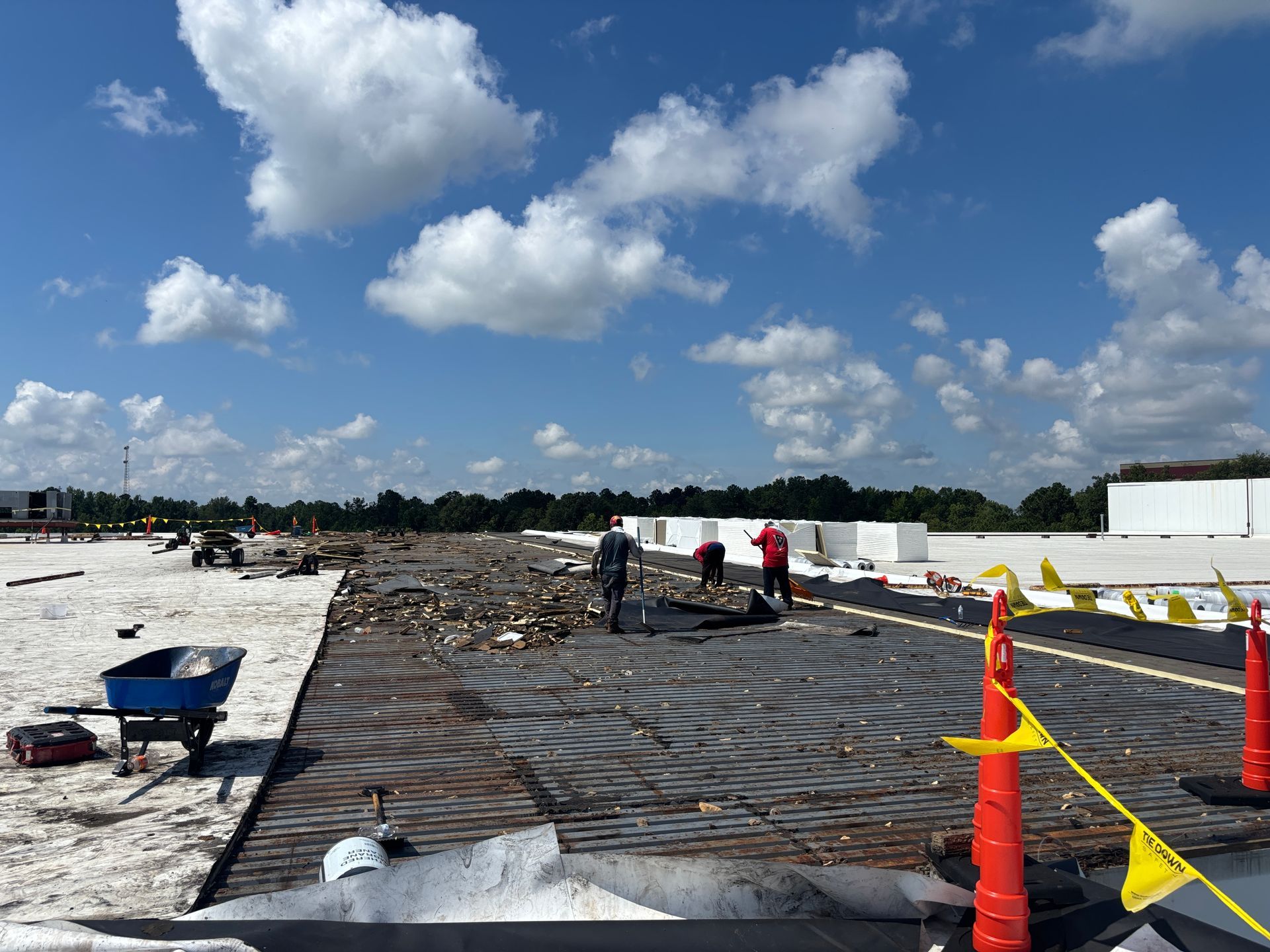 Workers on a commercial rooftop removing old roofing under a partly cloudy sky.