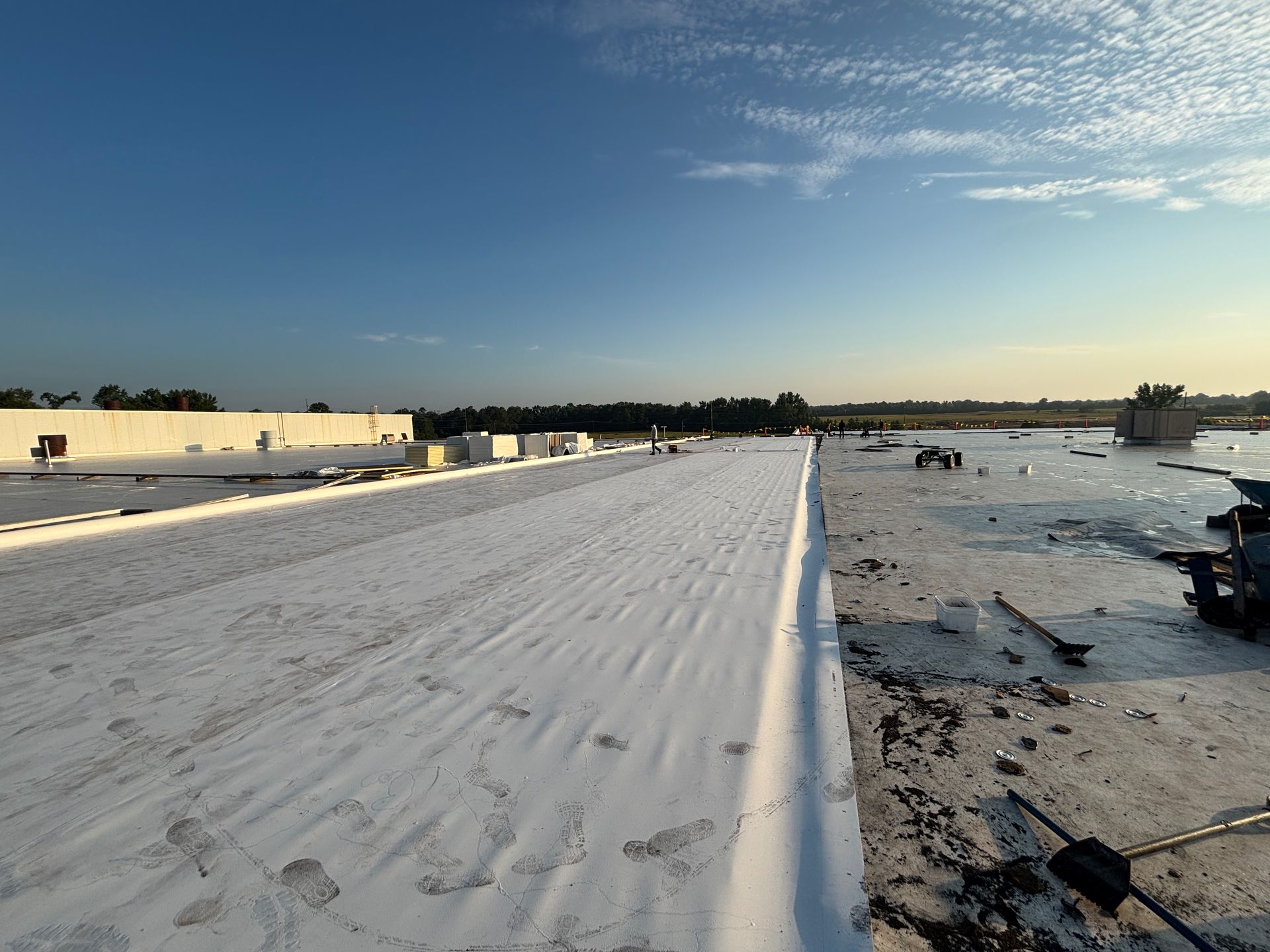 Flat commercial rooftop with white membrane installation, blue sky, and distant trees.