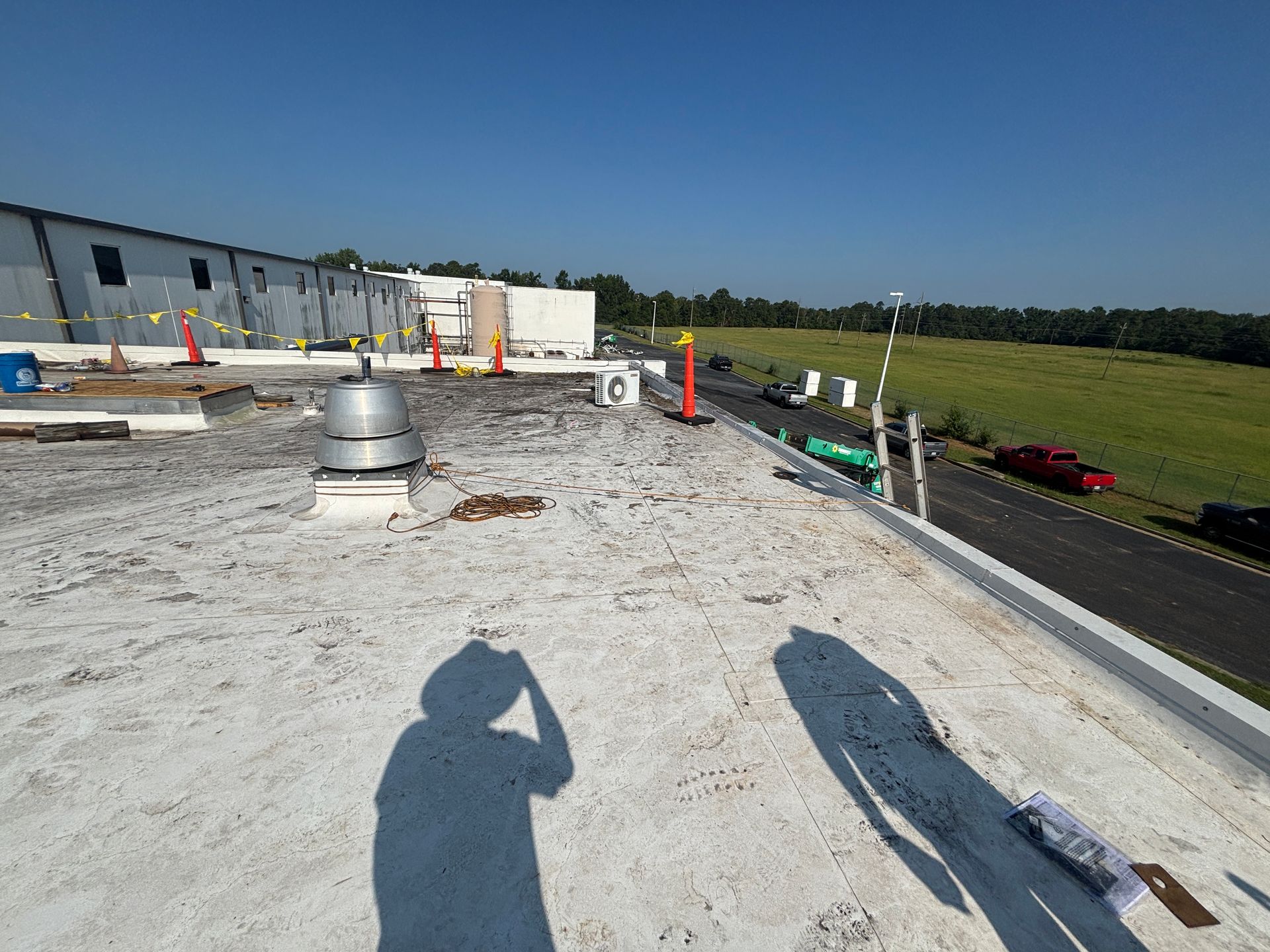 Shadow of person on a flat, white roof with vents.  Road and field in the background. Clear, blue sky.
