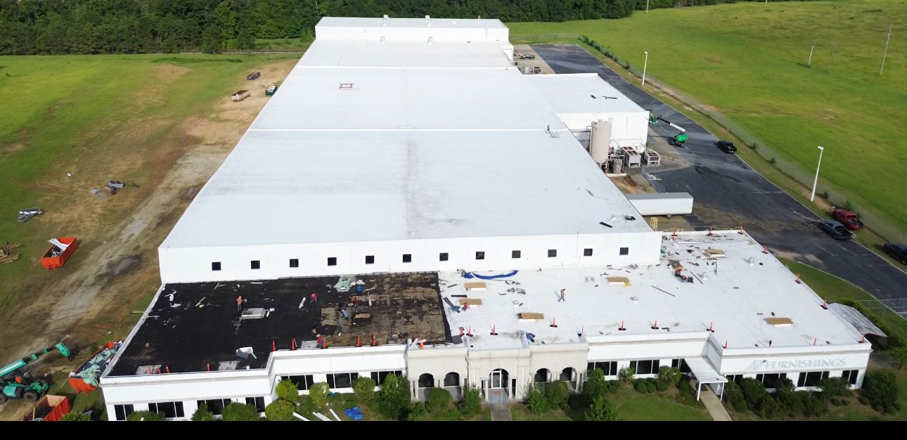Aerial view of a large white commercial building with damaged roof. Surrounding it is green grass and trees.