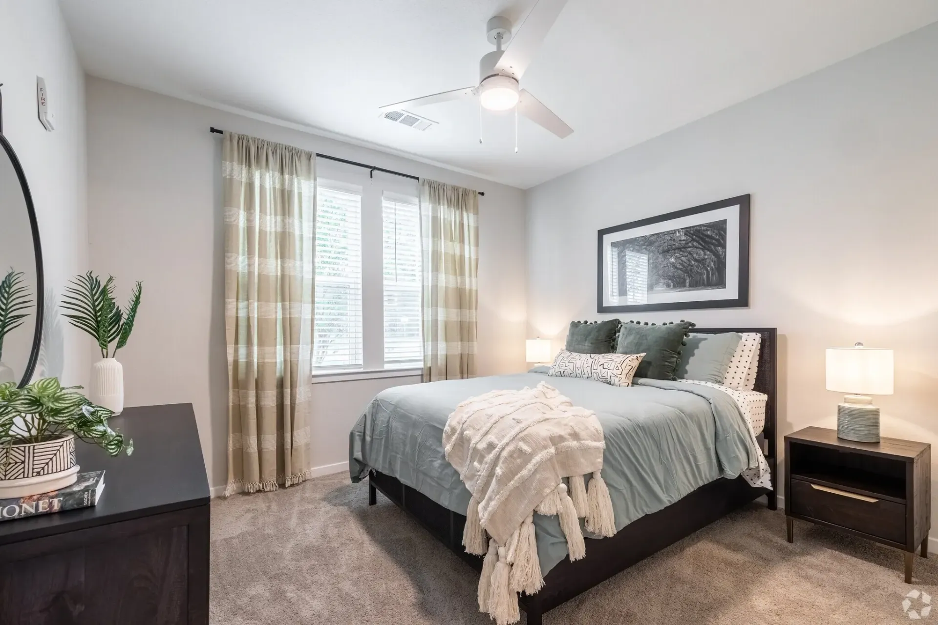 Bright bedroom with a gray bed, white walls, beige curtains, and bedside lamp near a window.