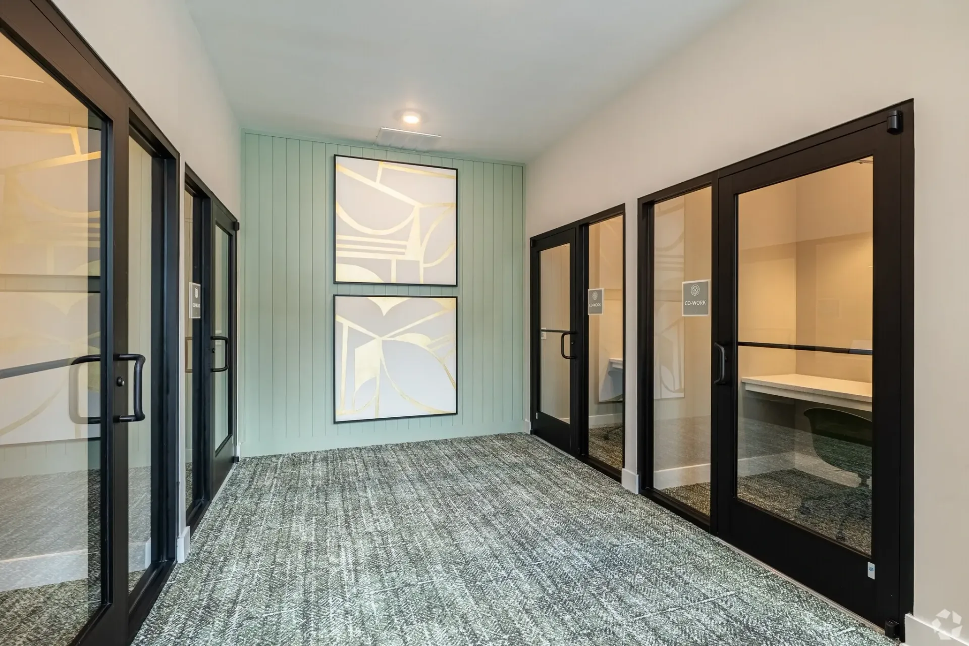 Hallway with glass-enclosed co-working spaces and abstract art on mint green wall.