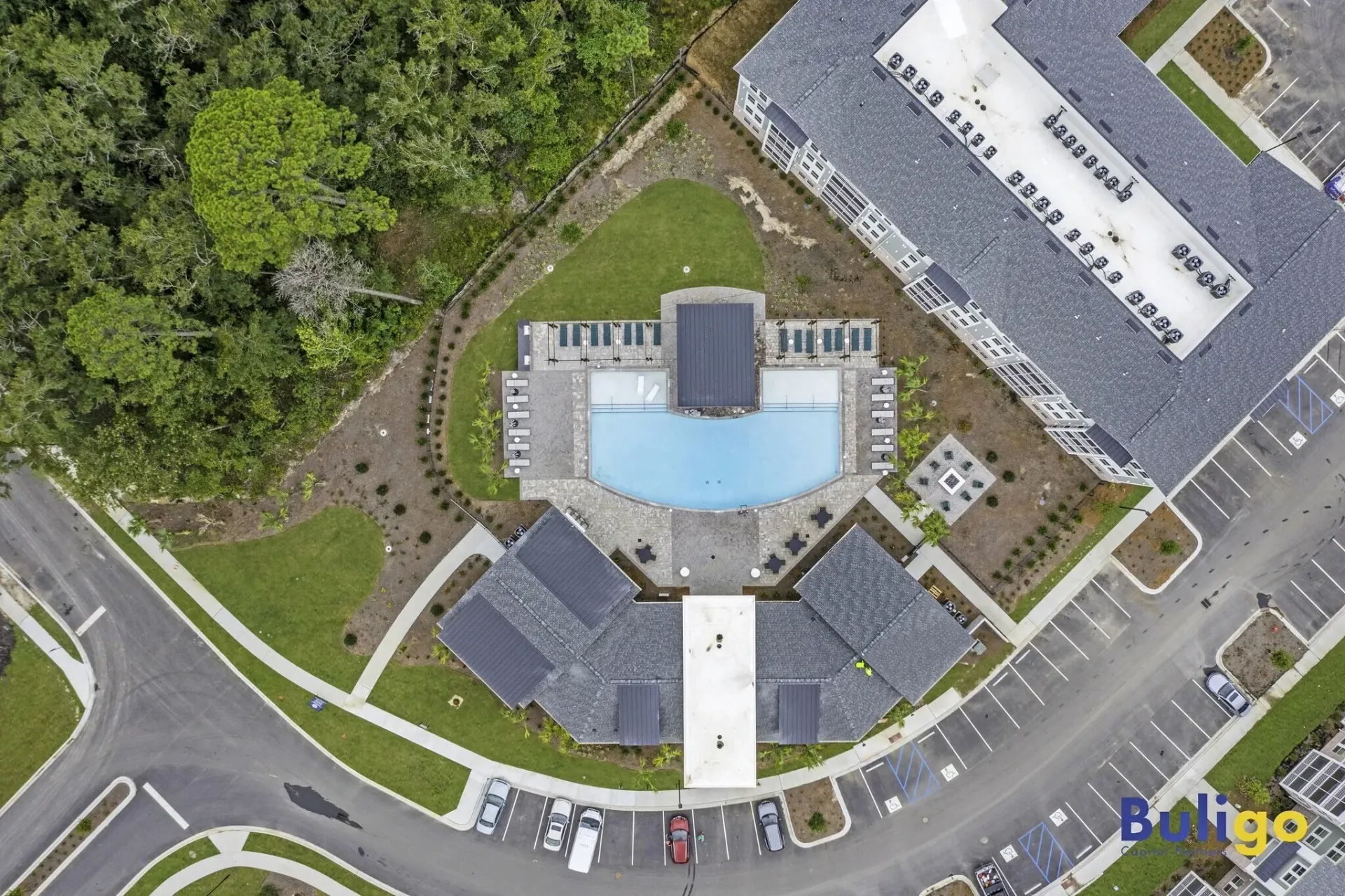 Aerial view of a modern apartment complex featuring a large outdoor swimming pool with lounge chairs, surrounded by buildings and parking lots.