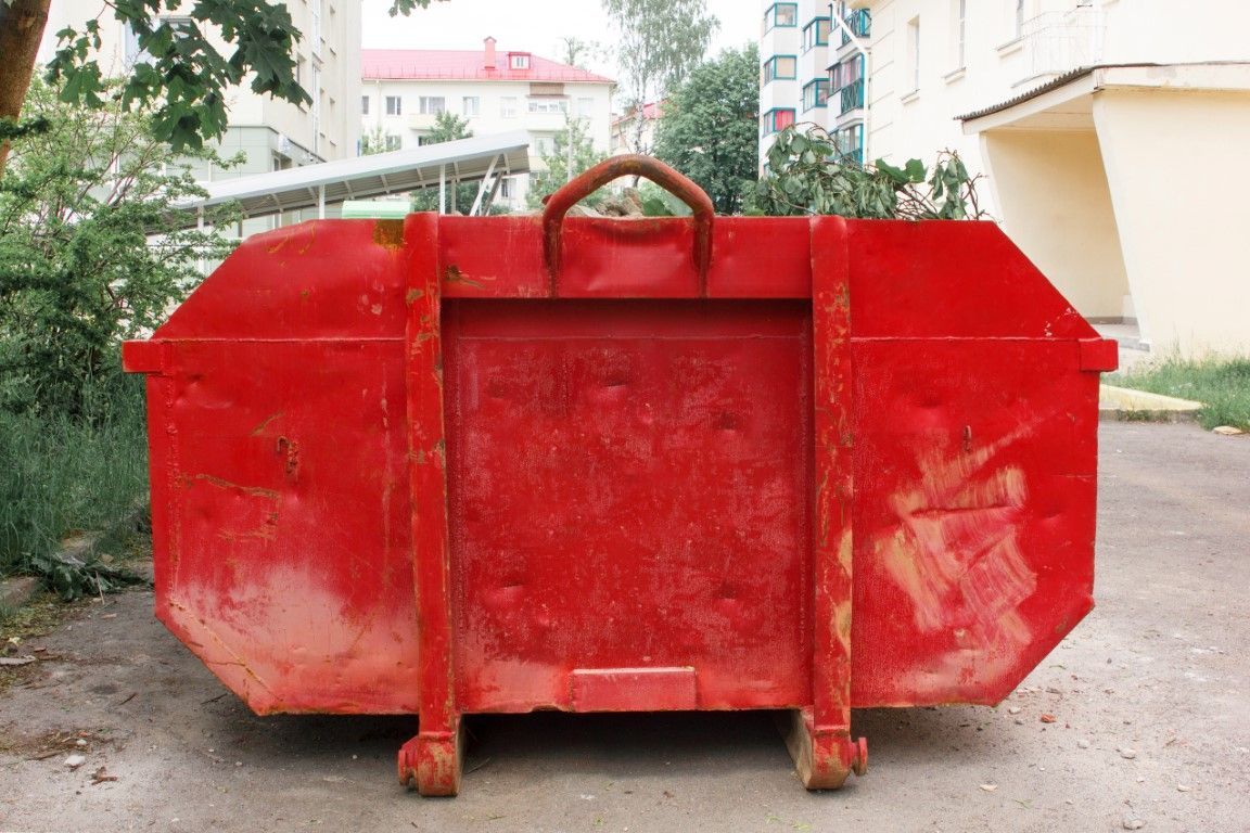 Red metal dumpster on a dirt path outdoors, with buildings and greenery in the background
