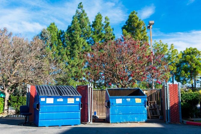 Two blue dumpsters beside a fence, with trees and a clear sky in the background.