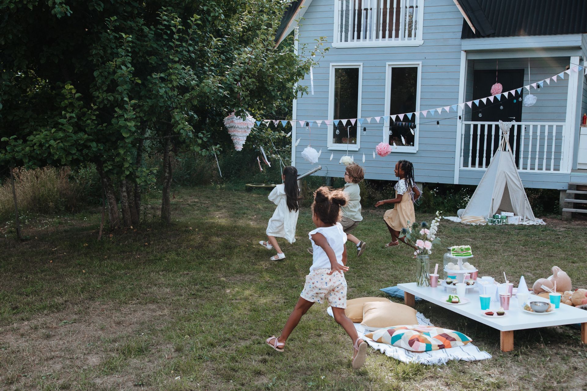 Children playing in a grassy backyard beside a blue house with a picnic table and teepee tents