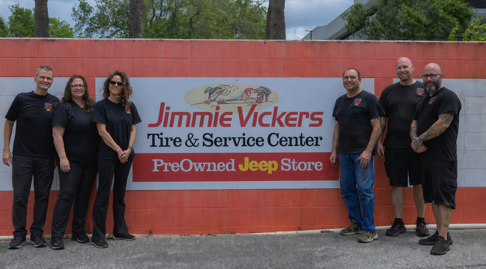 Six people posing outside Jimmie Vickers Tire & Service Center sign, beside a red wall.