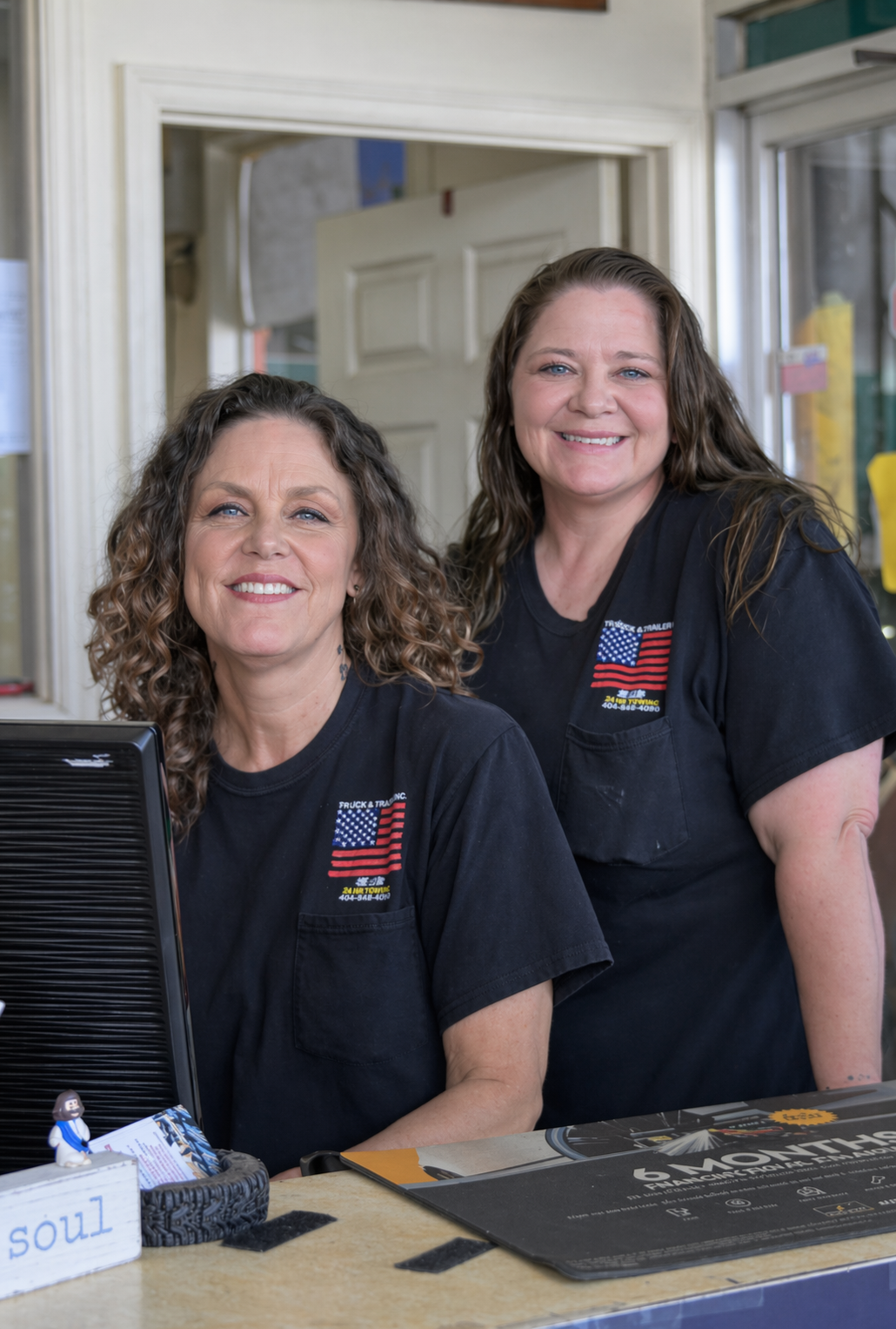 Two women in black shirts smiling behind a reception desk inside a building