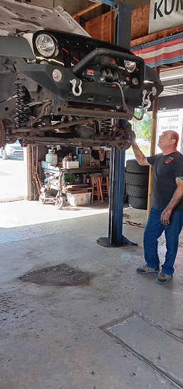 Man in blue overalls standing beside a car lifted on a hydraulic garage hoist