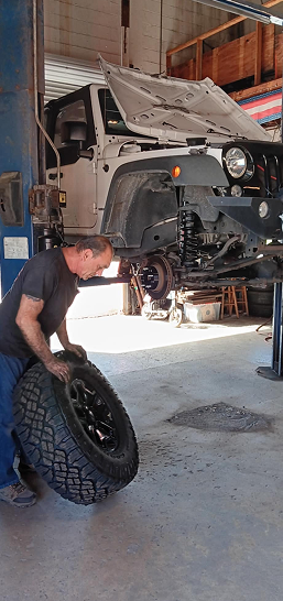Mechanic working on a lifted off-road vehicle in a garage, holding a large tire with the hood open