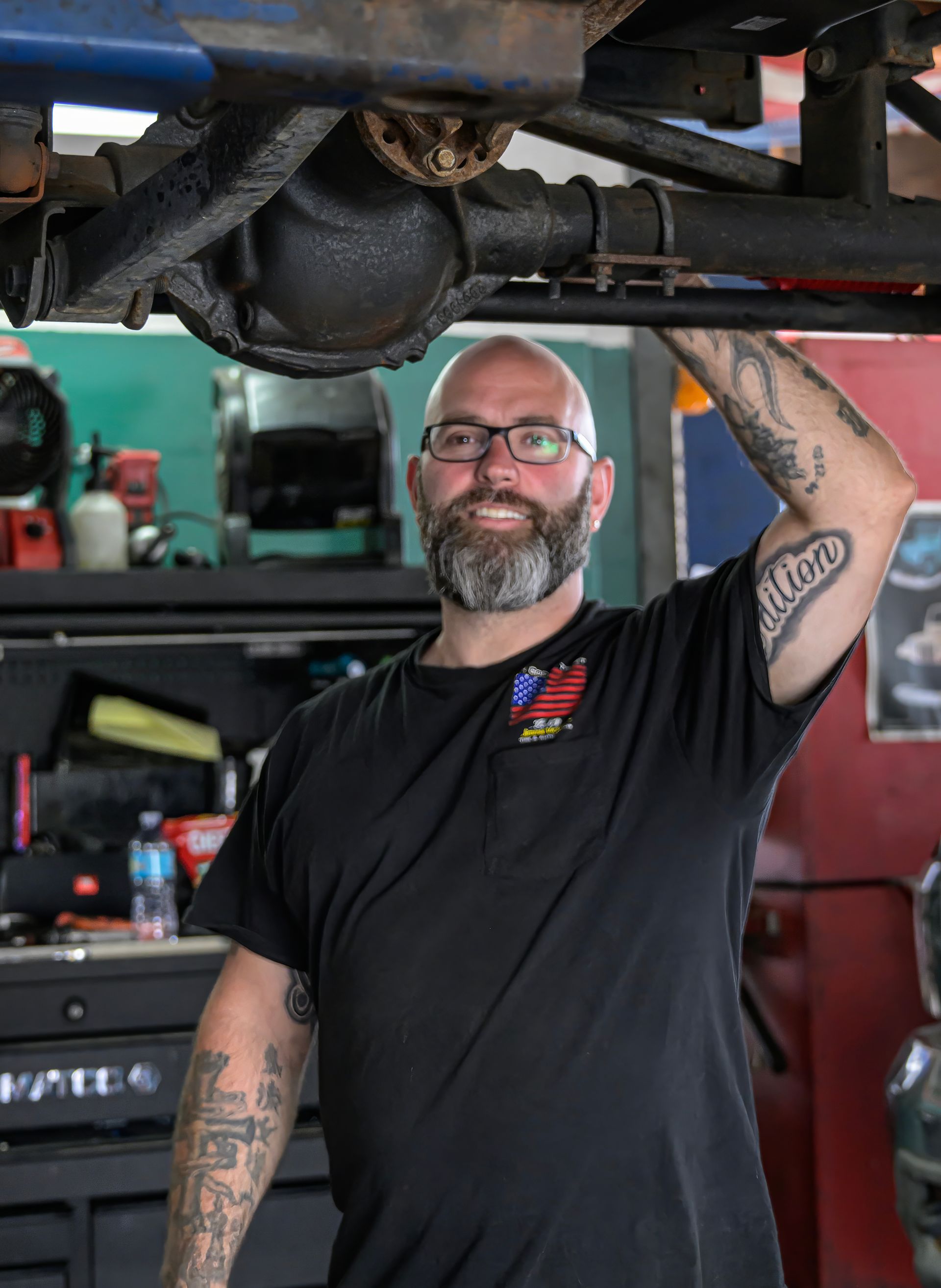 Mechanic smiling under a lifted vehicle in a garage, wearing a black shirt and glasses.
