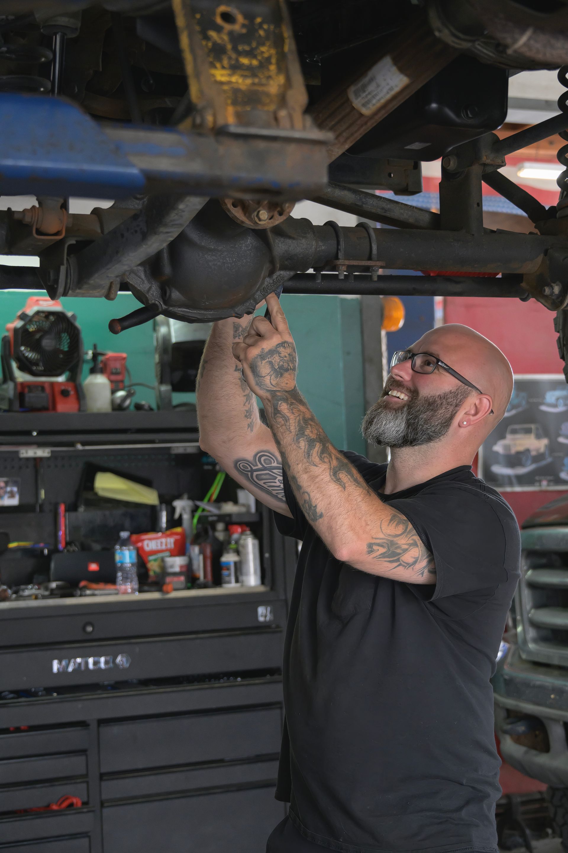 Mechanic working under a lifted vehicle in a garage, using a tool on the suspension.