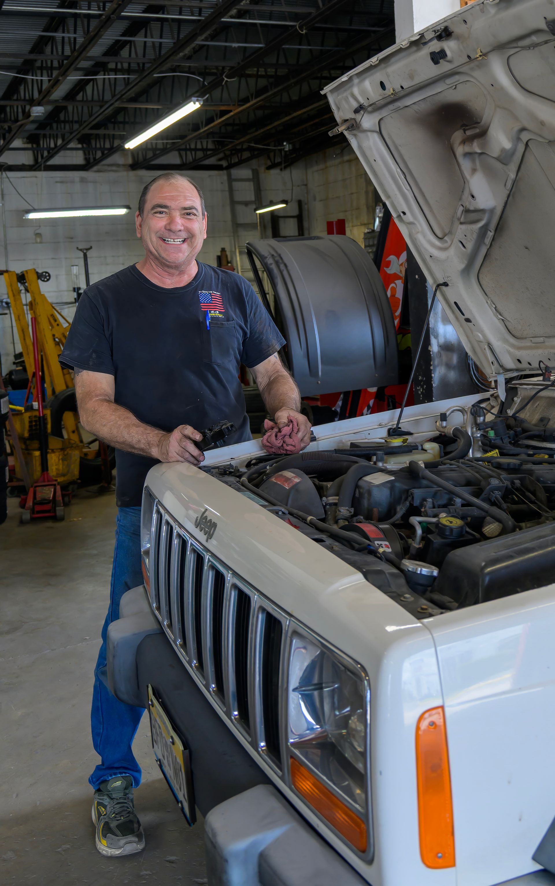 Mechanic smiling beside open hood of a white truck in a garage, holding tools