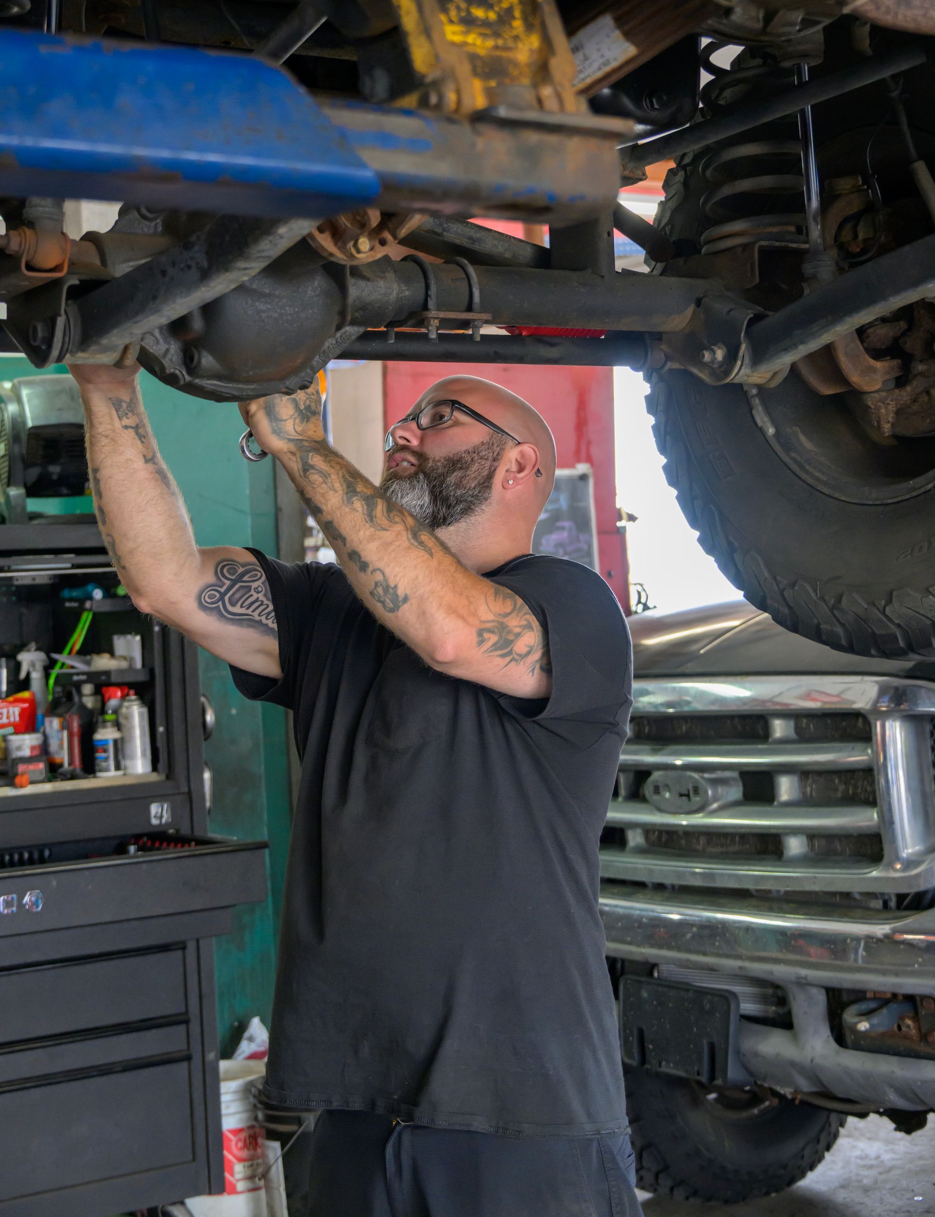Mechanic working under a lifted truck in a garage, using a tool on the undercarriage.