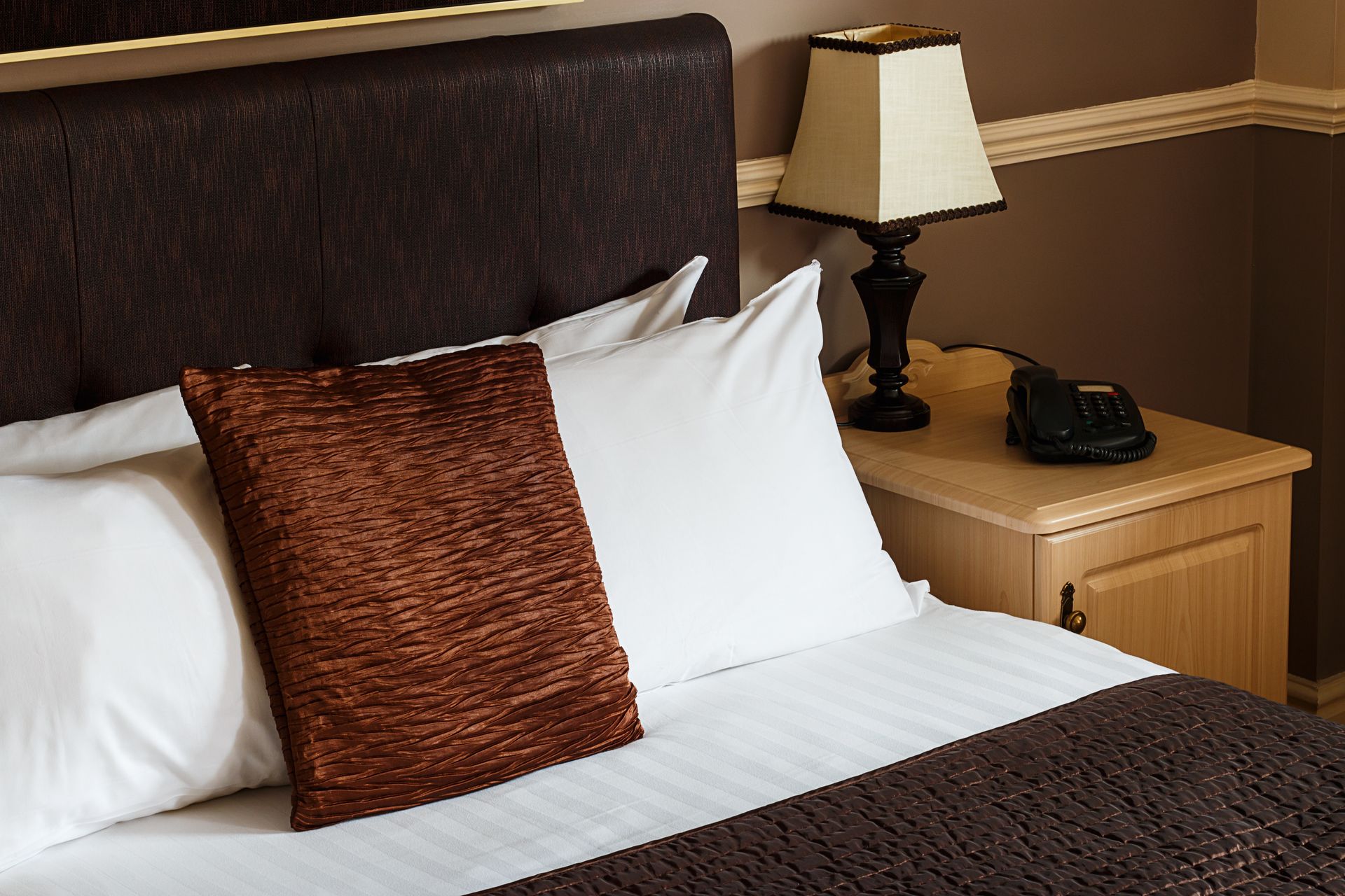 Bed with white sheets, brown headboard, decorative pillows, nightstand with lamp and phone.