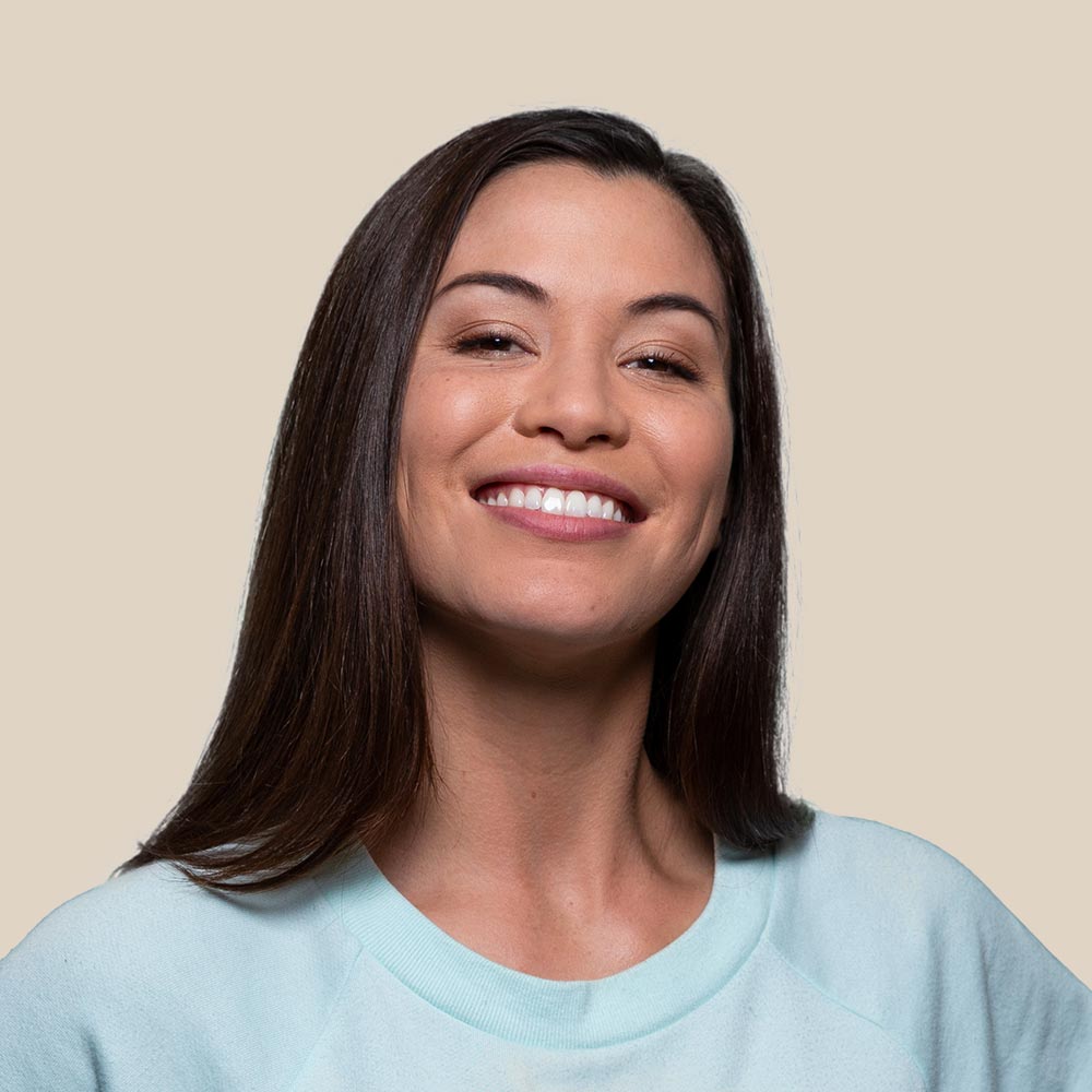 Woman with long dark hair, smiling in a light blue sweatshirt, against a beige backdrop.