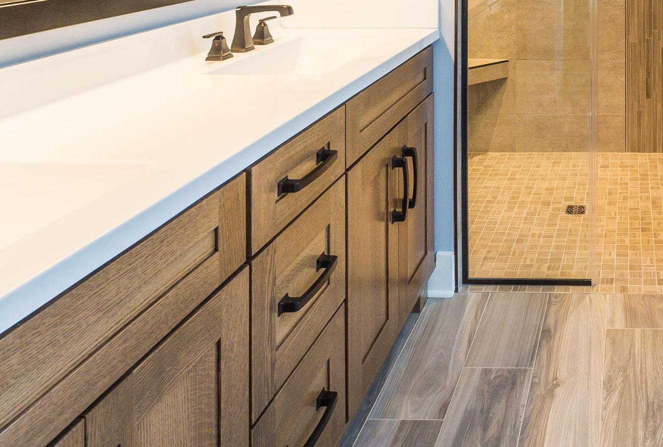 Bathroom vanity with light wood cabinets, white countertop, and black hardware. A shower is visible in the background.