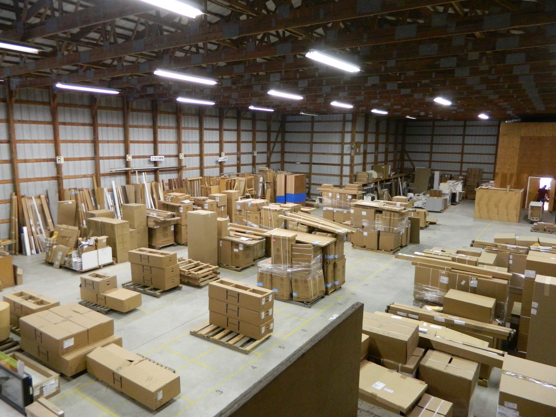 Warehouse interior with numerous cardboard boxes stacked on pallets and the floor, under bright overhead lighting.