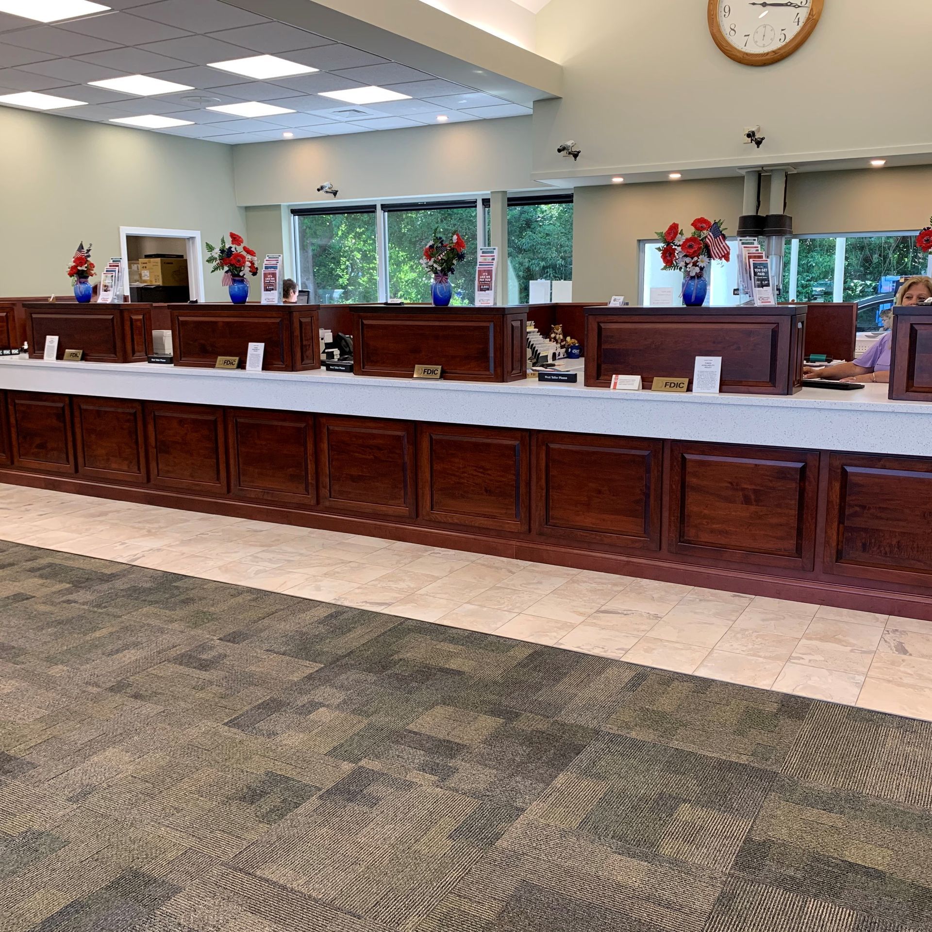 Inside a bank, a long, dark wooden counter with multiple teller stations is visible. Flowers and small signs decorate each station.
