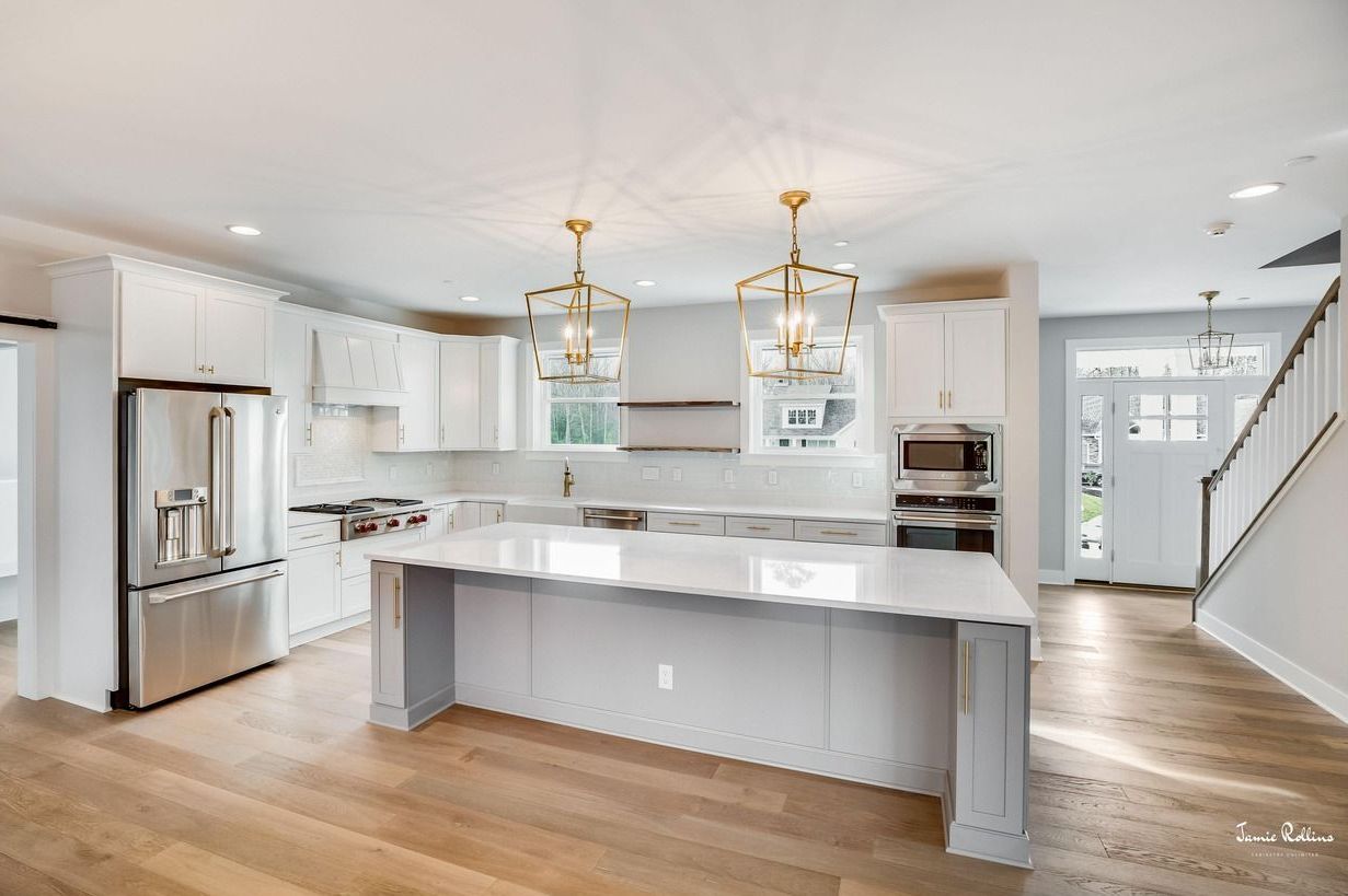 Modern, bright kitchen with white cabinets, island, and stainless steel appliances. Wooden floors, gold pendant lights, and a staircase are visible.