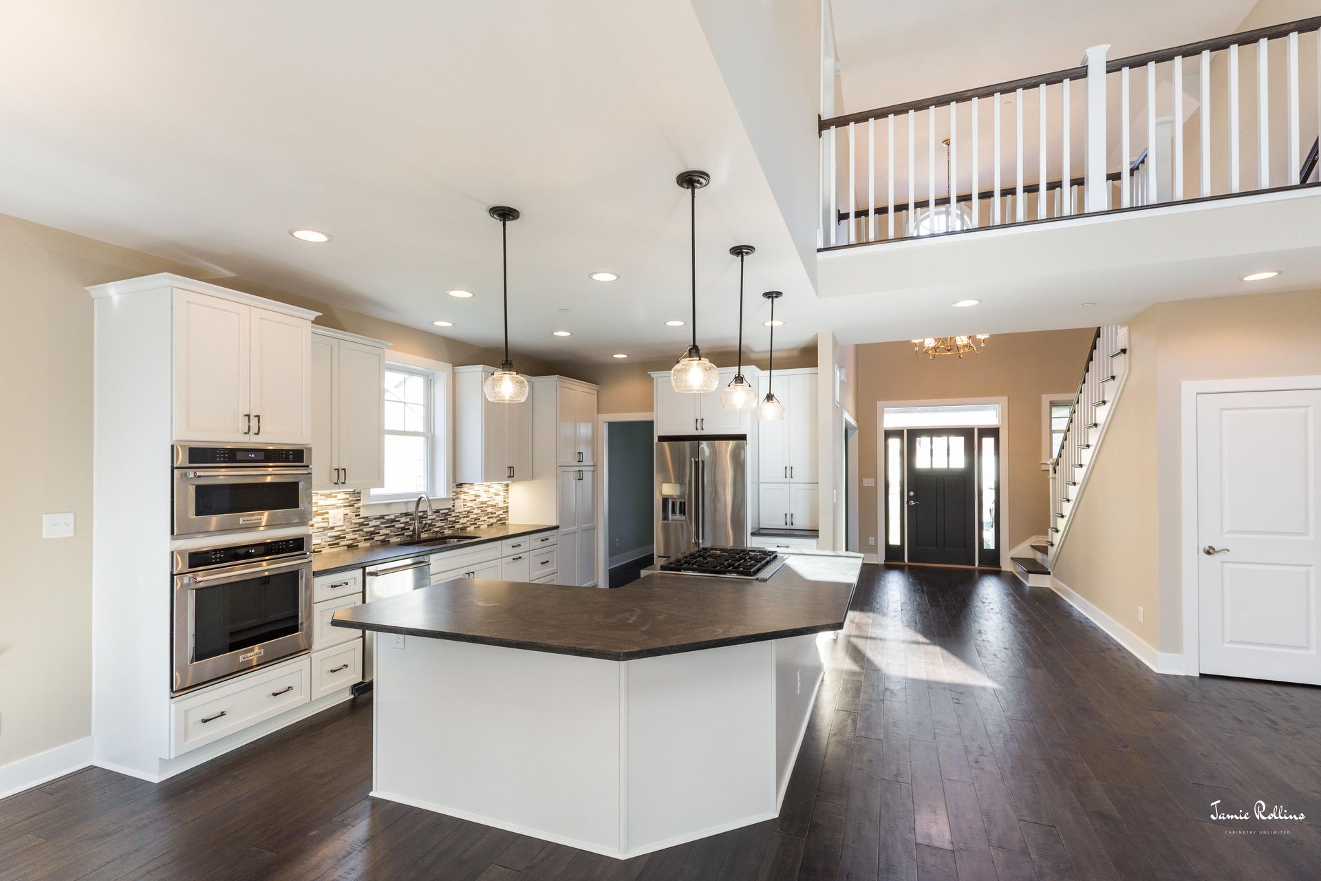 Open-concept kitchen with white cabinets, dark island countertop, stainless steel appliances, and a staircase leading to a loft.