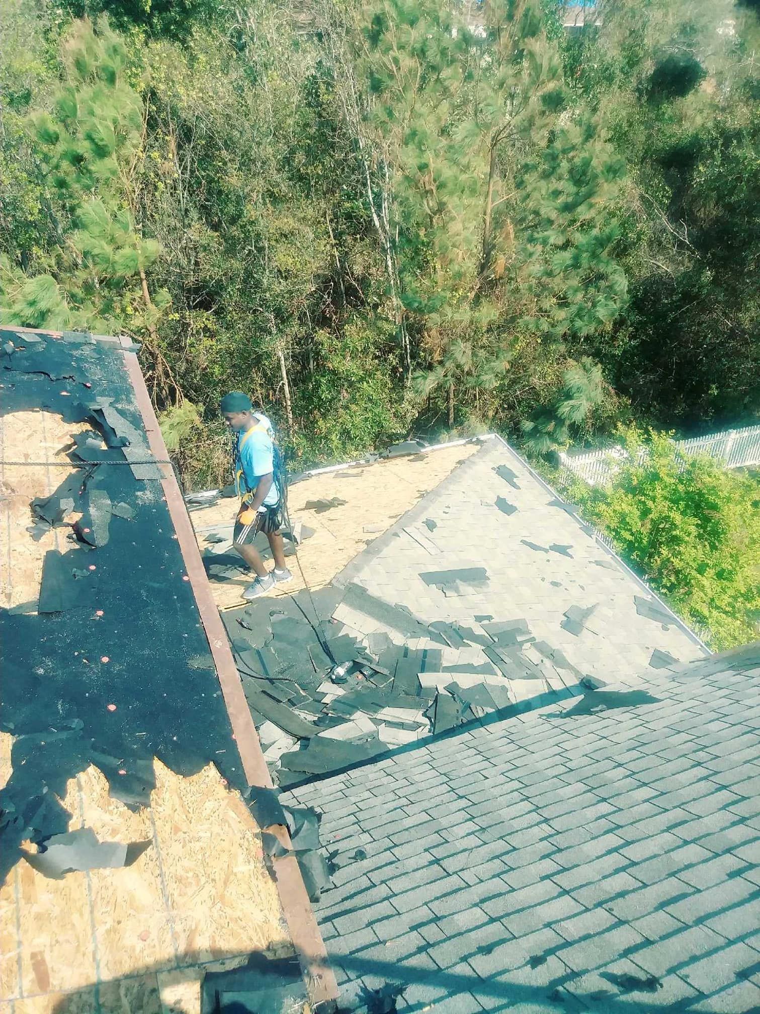 Roofer removing old shingles from a roof, set against a backdrop of green trees.