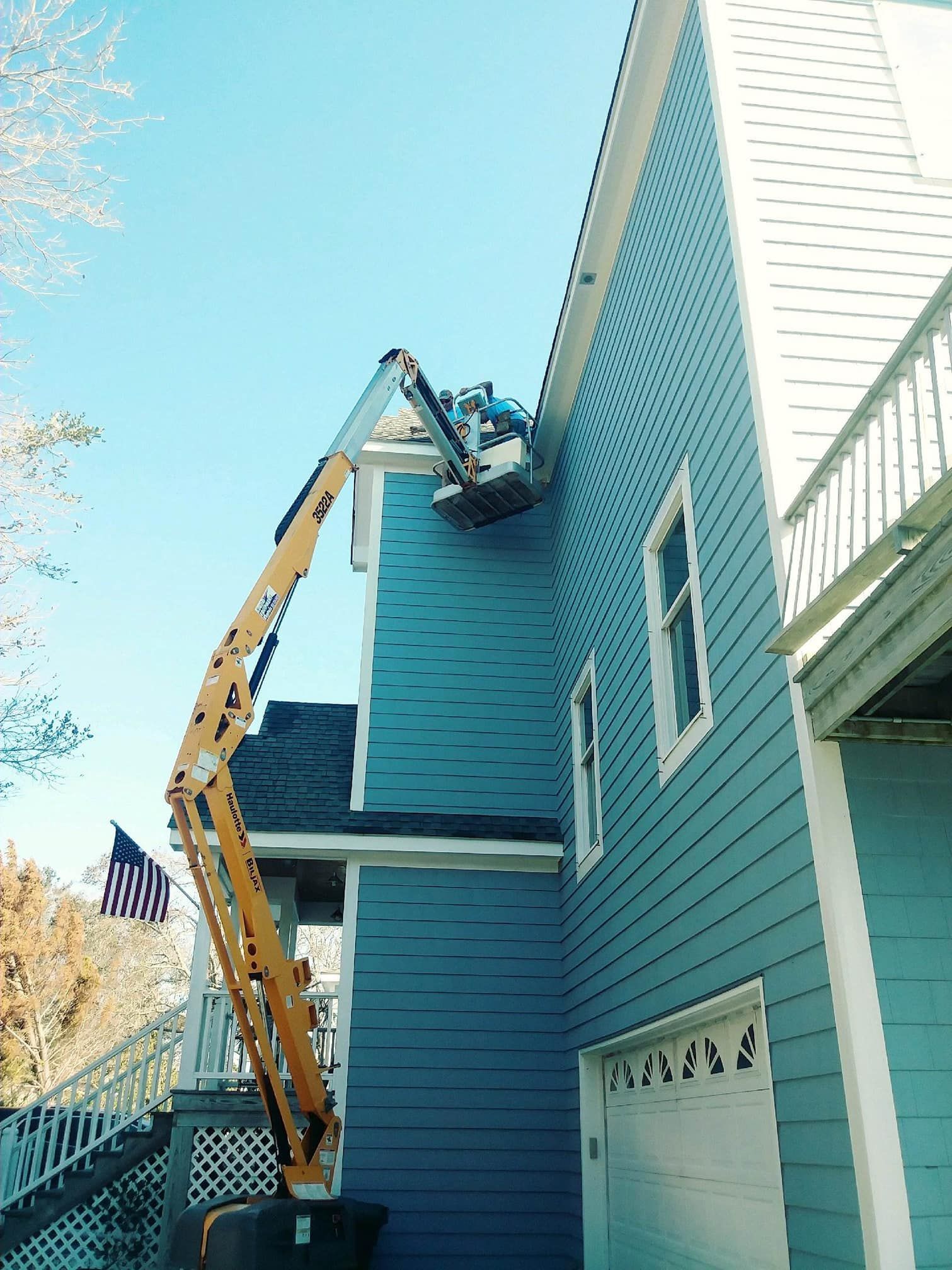 A cherry picker is next to a blue house, possibly repairing the roof. American flag waves.