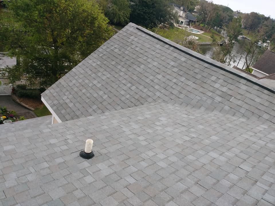 Gray shingle roof with a vent pipe, viewed from above. Background features trees and a lake.