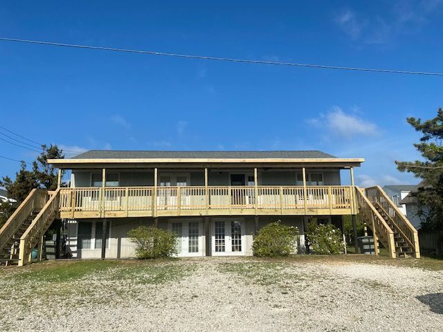 Two-story beach house with wooden decks, stairs, and white doors under a blue sky.