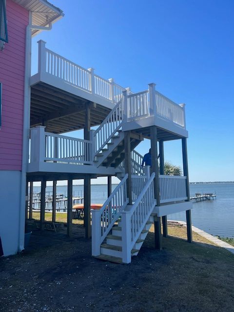 Three-level white deck with stairs connected to a pink house, overlooking a body of water under a blue sky.